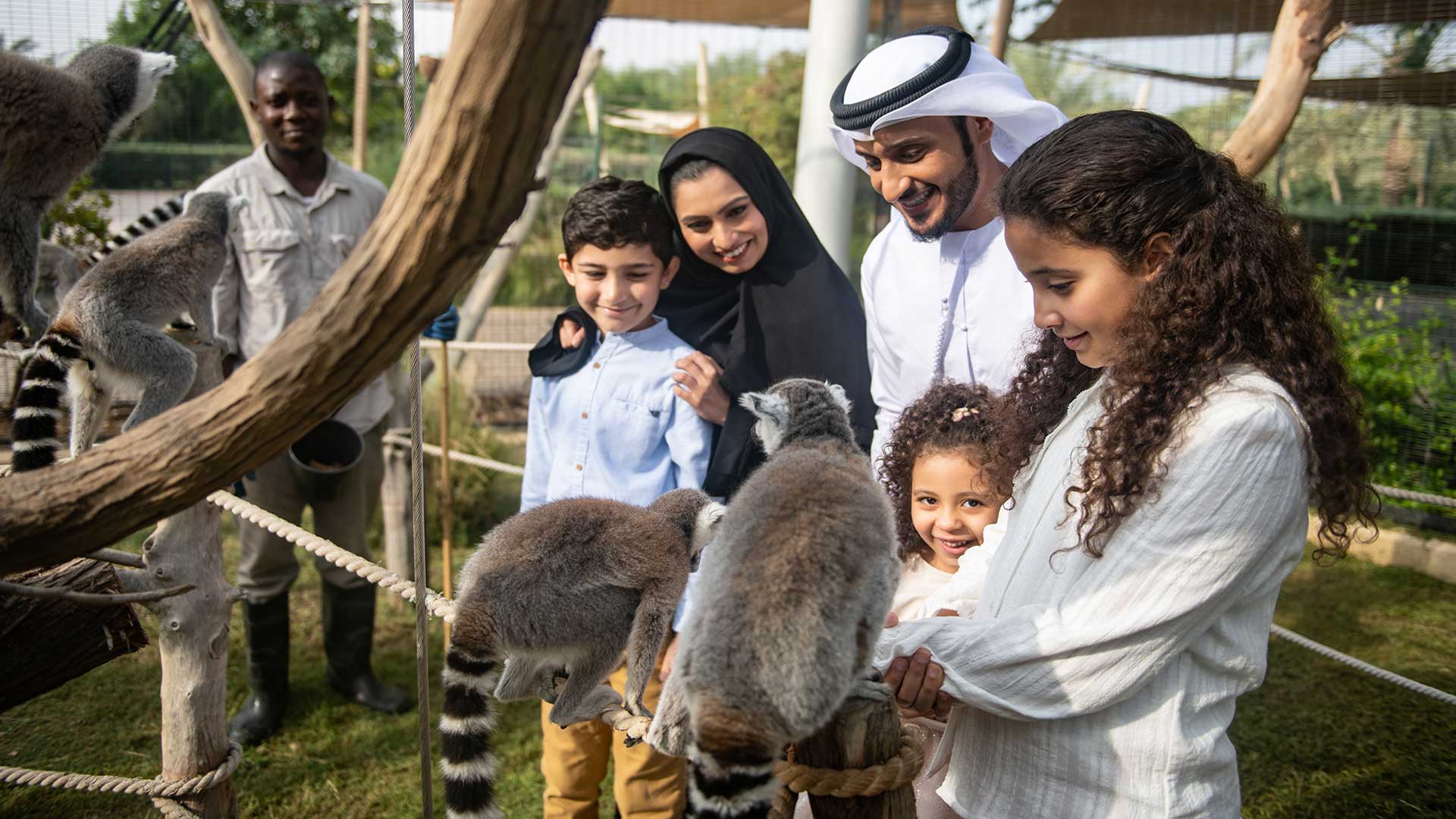 A family interacts with ring-tailed lemurs at an outdoor animal exhibit.