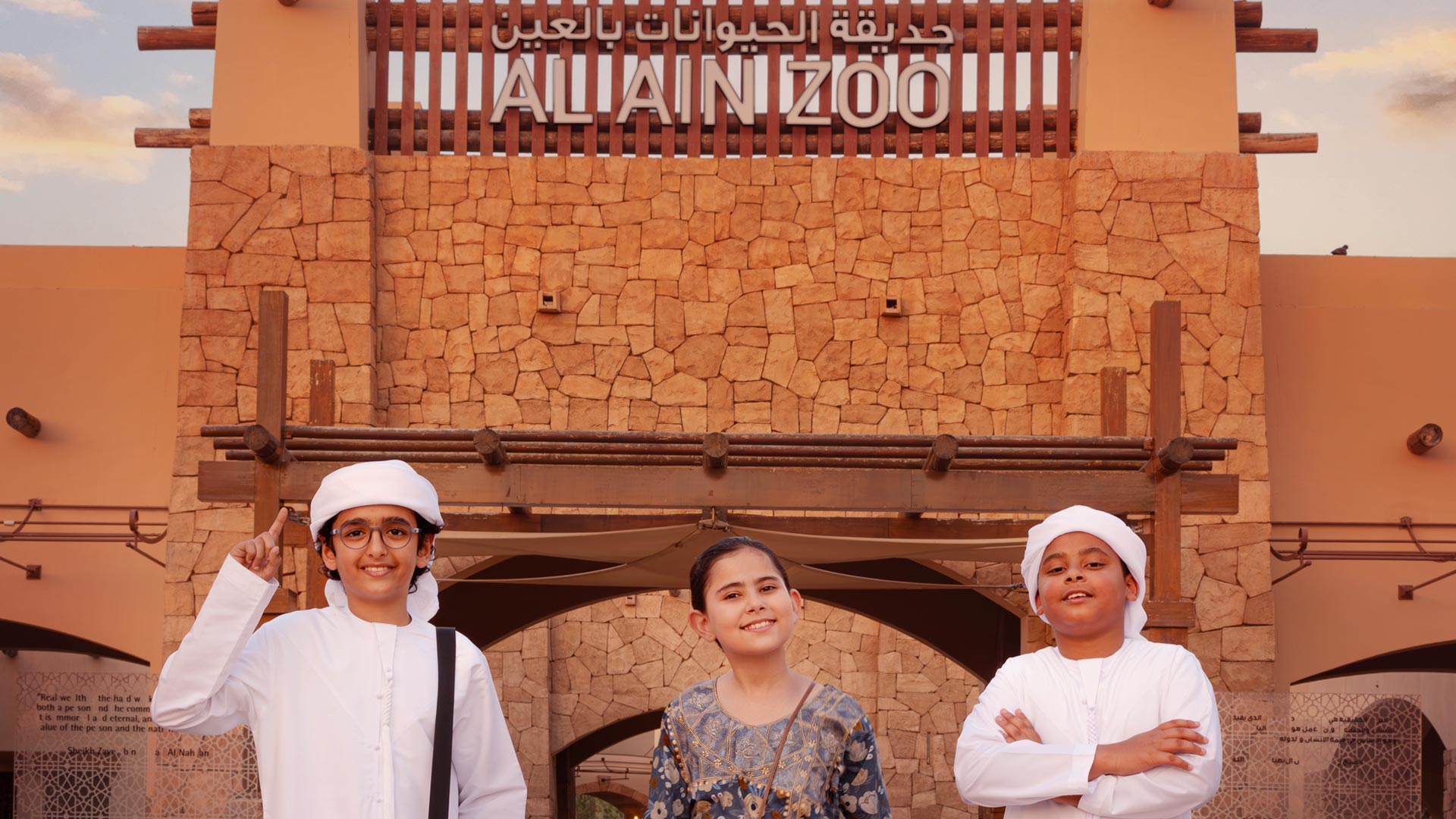 3 Emirati children dressed in traditional dress standing in front of Al Ain Zoo entrance