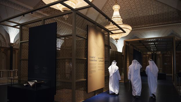 Three visitors in white clothing walk through an ornate museum exhibition beneath illuminated chandeliers