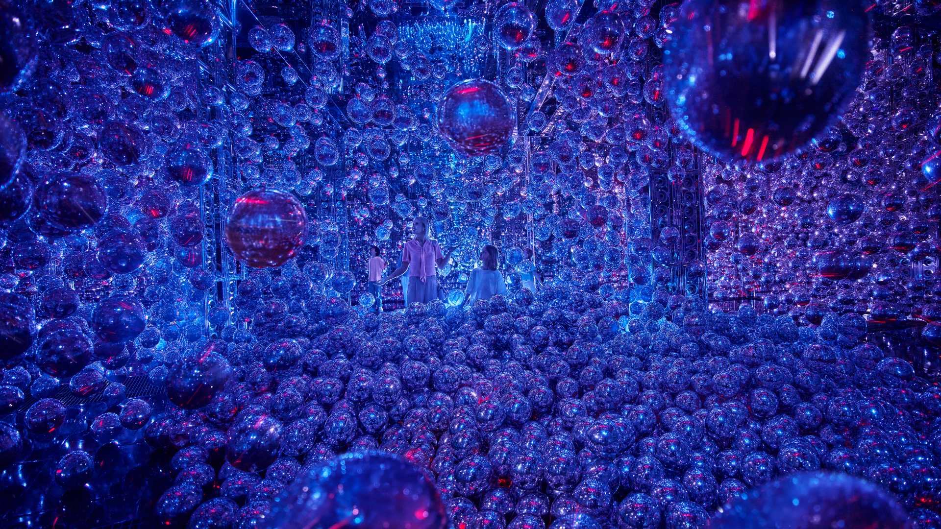 A mother with her son and daughting interacting with floating silver balloons with purple-blue hues in the exhibit.